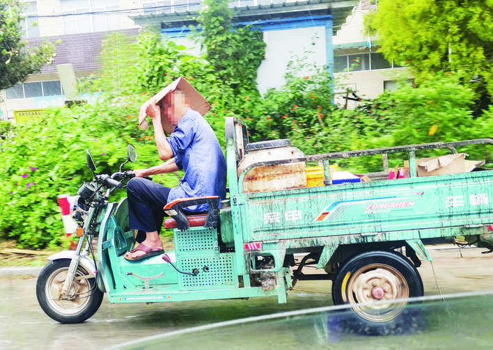 因下雨,一位大爷一手拿纸板遮雨,一手扶车把,开着三轮摩托车行驶在